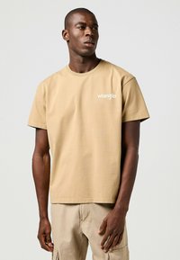 Camiseta de manga corta beige hecha de algodón con cuello redondo y un logo blanco de "Wrangler" en el pecho, presentada sobre un fondo neutro.