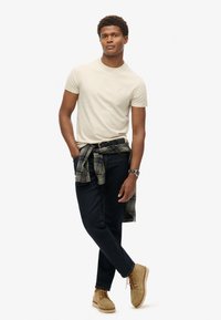 Beige t-shirt, mørke jeans, lysebrune sko. Grønternet skjorte bundet om livet. Yderligere detaljer inkluderer et armbåndsur og et bælte.