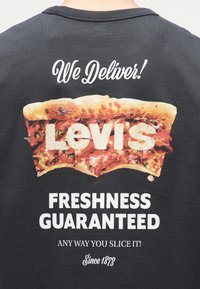 Μαύρο πουκάμισο με γραφικό μια φέτας πίτσας που δείχνει τη λέξη "Levi's" από τυρί, το κείμενο γράφει "Κουβαλάμε! Φρεσκάδα εγγυημένη με όποιο τρόπο κι αν την κόψεις! Από το 1873."