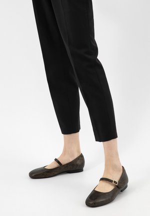 Persona che indossa pantaloni neri a gamba stretta e scarpe Mary Jane marrone scuro con texture e una piccola fibbia, su sfondo chiaro.