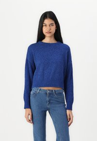 Suéter azul cortado em malha com um tecido texturizado, decote redondo e bainha e punhos com ribanas, combinado com jeans azuis ajustados.