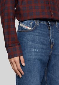 Keskitummansiniset farkut, joissa näkyvät tikkaukset; yhdistetty punaisen ja laivastonsinisen ruudullisen paidan kanssa. Takataskussa merkintä.