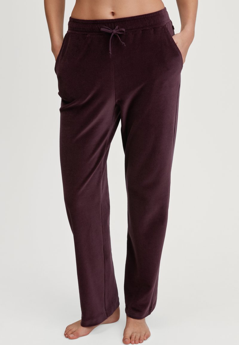 Persoon in donkerpaarse fluwelen broek met een trekkoord in de taille, die blootsvoets met de handen in de zakken tegen een effen achtergrond staat.