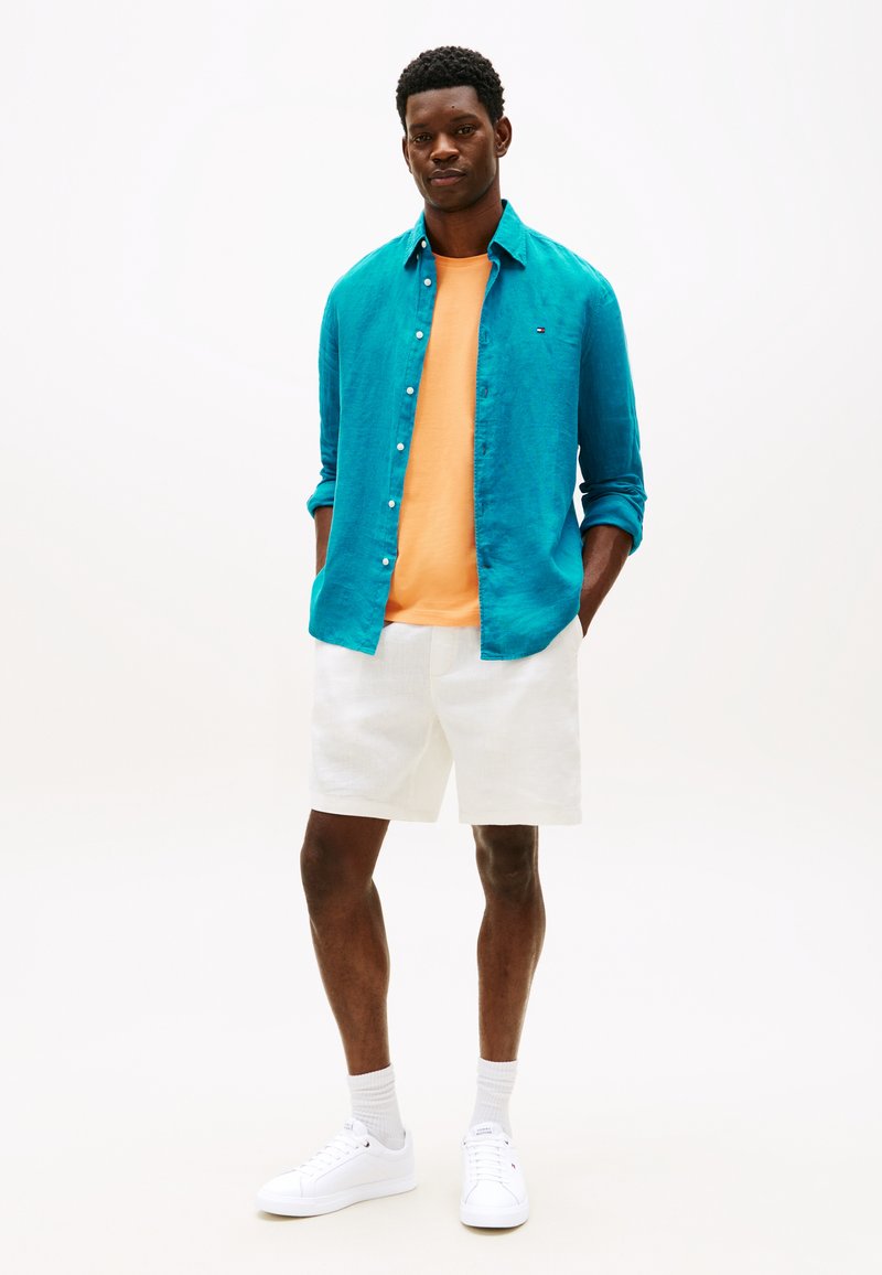 Τυρκουάζ πουκάμισο λίνεν με κουμπιά πάνω από πορτοκαλί μπλουζάκι, συνδυασμένο με λευκά σορτς. Το μοντέλο φοράει λευκά αθλητικά παπούτσια και γκρι κάλτσες, στέκεται μπροστά σε ανοιχτό φόντο.