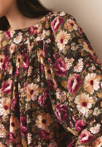 Bluză florală în culori verde închis, vișiniu și crem. Prezintă modele mari de flori, textură pliată și mâneci lungi, largi.