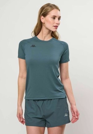 Žena v laděném modrozeleném sportovním tričku a krátkých kalhotách, dívající se doprava na jednoduchém světlém pozadí.