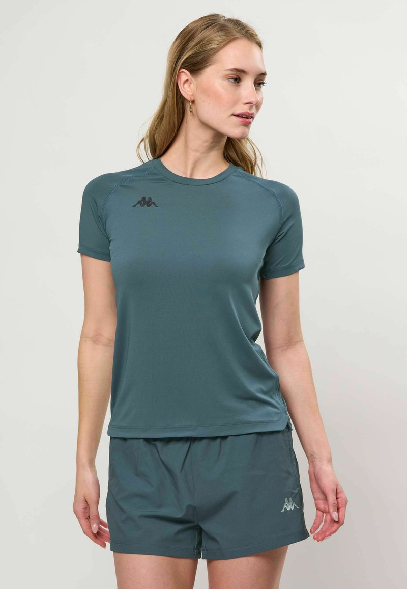Femme portant un t-shirt de sport et un short assortis de couleur bleu sarcelle, regardant vers sa droite sur un fond clair uni.