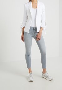 Blazer bianco in lino con maniche a cravatta, abbinato a jeans skinny azzurri chiari e sneakers bianche. Linee pulite e design minimalista.