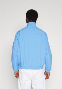 Chaqueta cortavientos azul claro con cuello alto, puños y dobladillo elásticos. Textura suave y corte relajado, sin patrones visibles ni elementos metálicos.