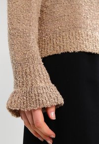 Pull en maille marron clair avec tissu texturé, poignets côtelés et coupe décontractée. La manche présente un ourlet évasé au niveau du poignet.