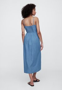 Duga haljina od trapera s naboranim stražnjim dijelom, tankim podesivim naramenicama i opuštenim krojem, u svijetloplavoj boji. Ima ravni rub i suptilnu teksturu.