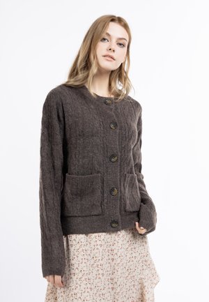 Junge Frau mit blondem Haar, die eine dunkelbraune gestrickte Strickjacke mit großen Knöpfen und einen hellen geblümten Rock vor weißem Hintergrund trägt.