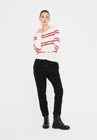Maglione bianco con righe orizzontali rosse e dettaglio del logo, abbinato a pantaloni neri a sigaretta e stivali neri stringati. Texture lisce.