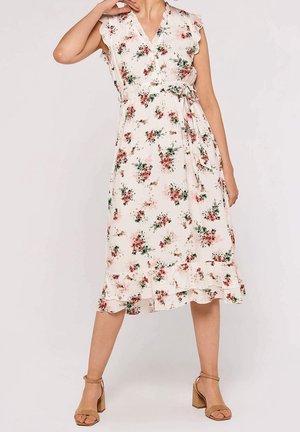 Mujer con vestido midi blanco sin mangas con estampado floral rojo y verde, cinturón atado a la cintura, bajo con volantes y sandalias camel con tacón cuadrado.