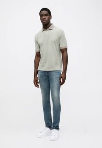 Világoszöld kötött galléros póló bézs gallérral és mandzsettával, fakó kék farmerral és fehér tornacipővel párosítva. Álló alak, elölről nézve.