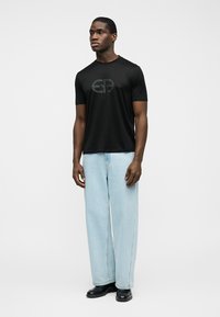 Άνδρας που στέκεται φορώντας μαύρο μπλουζάκι με λογότυπο, ανοιχτό μπλε παντελόνι με φαρδιές μπατζάκια και μαύρα παπούτσια, μπροστά σε απλό γκρι φόντο.