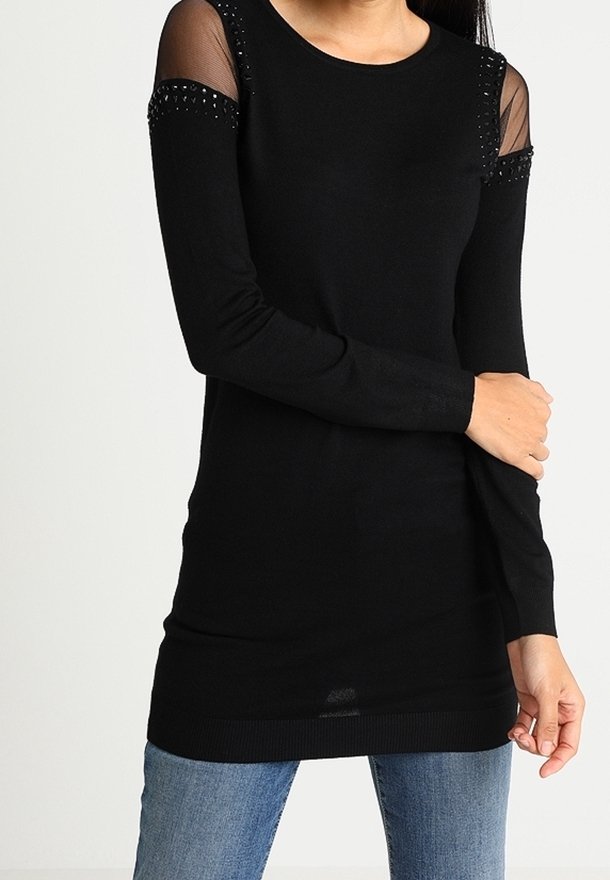 Femme portant un haut noir à manches longues avec découpes transparentes aux épaules et bordure cloutée, assorti à un jean bleu, posant devant un fond uni.