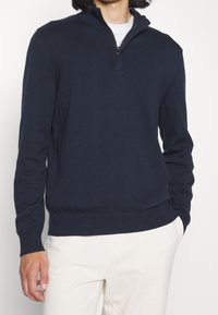Pull-over en maille bleu marine avec col zippé, poignets et ourlet côtelés, présentant une texture lisse et une coupe ajustée.