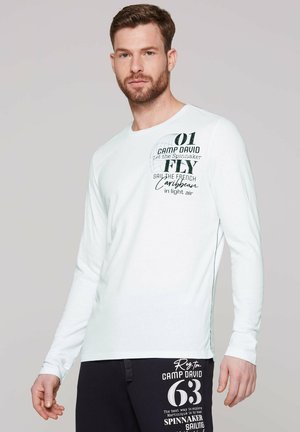Mann mit kurzen braunen Haaren und Bart, der ein weißes Langarmshirt und schwarze Hosen mit aufgedrucktem Text und Grafiken trägt.