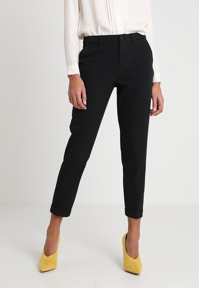 Femme portant un pantalon noir ajusté longueur cheville avec revers, un chemisier blanc à manches longues et des talons hauts jaunes pointus.