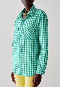 Camicia a scacchi verde e bianca con colletto, due tasche sul petto e chiusura a bottoni; indossata su pantaloni gialli con orlo sfrangiato.