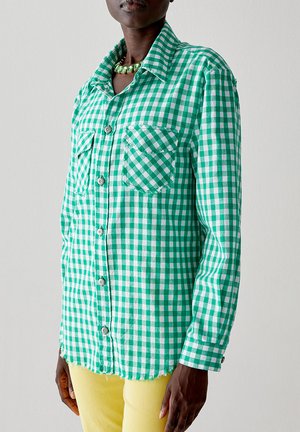 Camicia a scacchi verde e bianca con colletto, due tasche sul petto e chiusura a bottoni; indossata su pantaloni gialli con orlo sfrangiato.