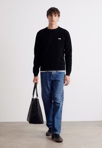 Czarna bluza z małym białym logo, niebieskie dżinsy oraz czarne obuwie. Trzyma czarną torbę tote na jasnym tle.