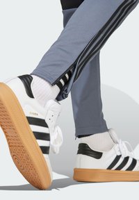 Weiße Sneaker aus Leder mit schwarzen Streifen, Gummisohle und Schnürdesign. Graue Sporthosen mit schwarzen seitlichen Reißverschlüssen. Weiße gerippte Socken.