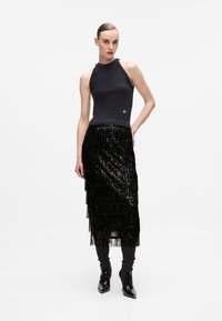 Haut noir sans manches avec col haut ; jupe noire à franges pailletées, longueur genoux. Le mannequin porte des bottes noires ajustées jusqu'à la cheville, mettant en valeur une silhouette élégante.