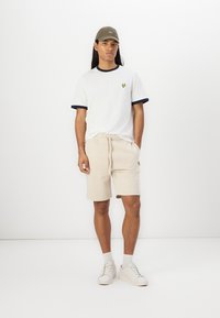 Creme shorts og hvid T-shirt med marineblå detaljer, udstyret med et lille gult logo. Sammen med en beige kasket og hvide sneakers.