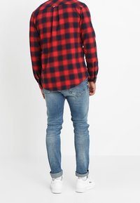 Chemise à carreaux rouge et bleu marine avec un col boutonné, des manches longues et un ourlet arrondi. Portée avec un jean délavé clair et des baskets blanches.
