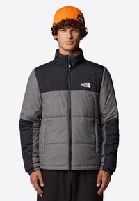 Pufferjack in grijs en zwart, met een hoge kraag, ritssluiting en elastische manchetten. Geweldig met een oranje geribde beanie.