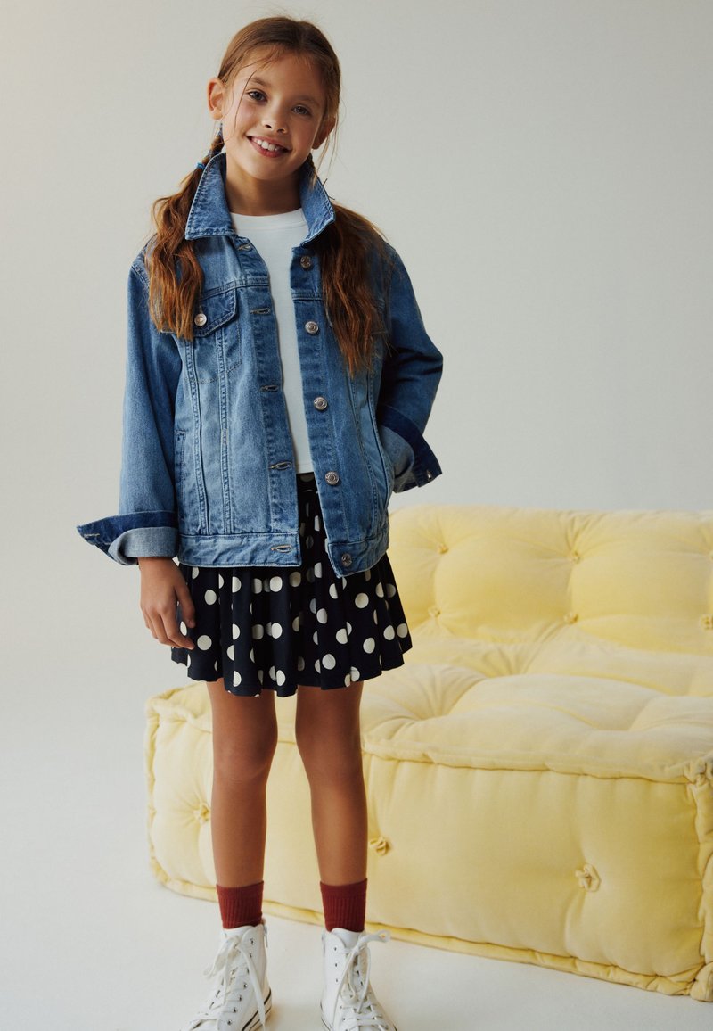 Smilende pige iført en denimjakke, hvid skjorte, sort nederdel med hvide prikker, røde sokker og hvide sneakers står ved siden af en gul sofa.