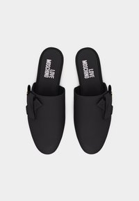 Ζευγάρι μαύρα παπούτσια χωρίς κορδόνια με φιόγκους στο πλάι και την επιγραφή "LOVE MOSCHINO" σε λευκό χρώμα στις εσωτερικές σόλες, απεικονιζόμενα από πάνω σε λευκό φόντο.