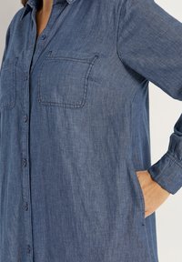 Tummansininen farkkupaita, jossa napit edessä, kaksi rintataskua, pitkät hihat ja löysä malli. Pehmeä rakenne ja rento muotoilu.