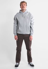 Sweat à capuche gris avec une poche avant et un accent à motif, associé à un pantalon marron et des baskets noires et blanches. Design simple et coupe décontractée.