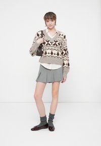 Une jeune personne se tient debout, vêtue d'un pull beige à motifs, d'une chemise blanche, d'une jupe plissée grise, de chaussettes grises, de chaussures bordeaux et portant un sac à bandoulière marron.