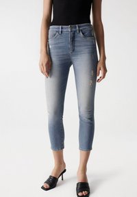 Jeans de mezclilla azul claro con un ajuste ceñido, que presentan detalles desgastados y una longitud corta, combinados con tacones abiertos negros.