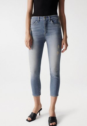 Jeans de mezclilla azul claro con un ajuste ceñido, que presentan detalles desgastados y una longitud corta, combinados con tacones abiertos negros.