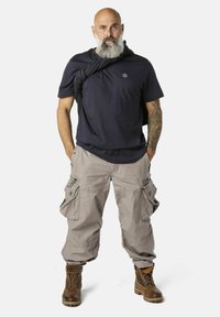 Dunkelblauses T-Shirt, Cargo-Hosen in Hellgrau mit mehreren Taschen und braunen Stiefeln. Ein Mann steht mit verschränkten Armen und neutralem Ausdruck.