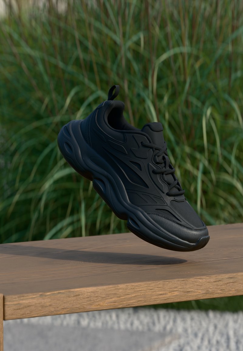 Μαύρο αθλητικό παπούτσι με υφασμάτινο επάνω μέρος με υφή, καμπυλωτή σόλα και διακριτικές σχεδιαστικές λεπτομέρειες. Εμφανίζεται στον αέρα πάνω από μια ξύλινη επιφάνεια.