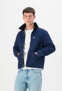 Chaqueta navy de cremallera con un collar negro texturizado, dos bolsillos laterales y un bolsillo en el pecho. Llevada sobre una camiseta blanca y unos vaqueros azul claro.