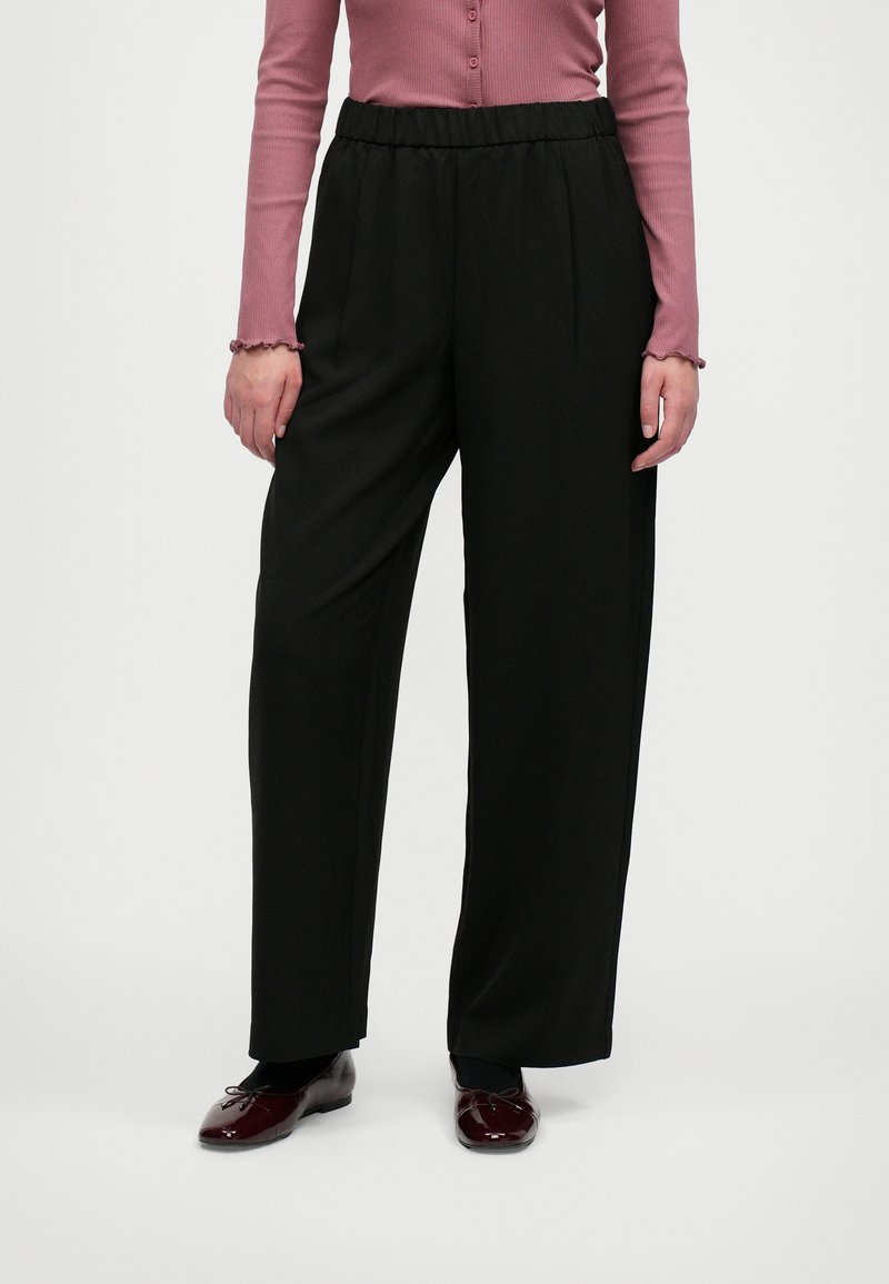 Pantalons larges noirs avec une taille élastique et des plis à l'avant. Portés avec un haut ajust�é à manches longues rose clair côtelé et des chaussures bordeaux.