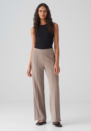 Tan wijde broek met verticale naden, gecombineerd met een strakke zwarte tanktop. De stof lijkt glad met een lichte glans.