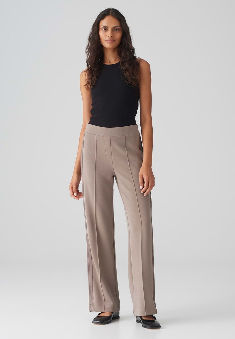 Tan wijde broek met verticale naden, gecombineerd met een strakke zwarte tanktop. De stof lijkt glad met een lichte glans.