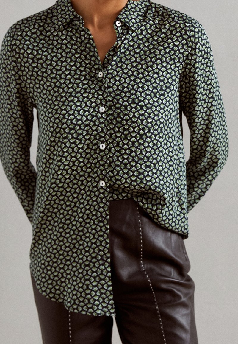 Personne portant une chemise noire à manches longues boutonnée avec un petit motif floral vert et blanc et un pantalon en cuir marron foncé avec des coutures blanches.