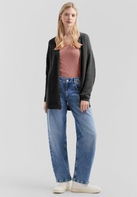 Młoda kobieta ubrana w ciemnoszary sweter dzianinowy, pudroworóżową tank top, luźne niebieskie dżinsy i białe sneakersy na jasnoszarym tle.
