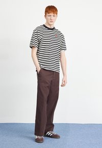 T-shirt à manches courtes rayé noir et blanc, pantalon décontracté marron foncé, et baskets marron avec accents et lacets blancs.