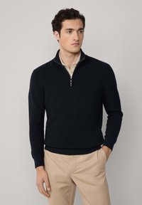 Man i en svart halvdragkedjesweater över en beige skjorta med krage och beige byxor, stående med en hand i fickan mot en enfärgad bakgrund.