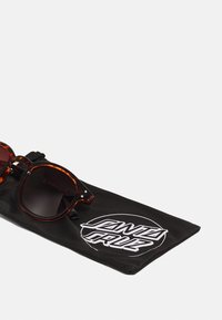 Sonnenbrille aus Schildpatt mit dunklen Gläsern, begleitet von einem schwarzen Beutel mit einem weißen "Santa Cruz"-Logo.